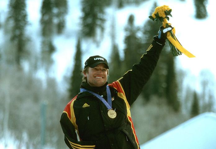A male athlete wearing a black uniform and a gold medal is celebrating, holding a flower bouquet with his left hand